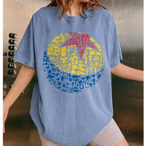 May include: A light blue short-sleeved t-shirt featuring a large circular design. The design is composed of a red star, yellow circle, and blue crescent, all filled with various cartoon character silhouettes. The shirt has a relaxed fit.