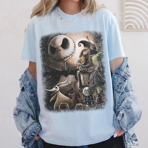May include: Light blue t-shirt featuring a graphic of characters from The Nightmare Before Christmas. The design includes Jack Skellington, Sally, Zero, and other characters in a dark, spooky scene. The shirt is a short-sleeved crew neck.