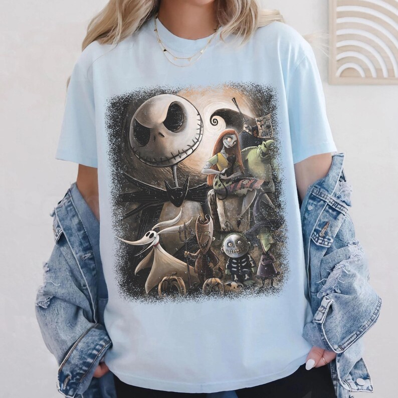 May include: Light blue t-shirt featuring a graphic of characters from The Nightmare Before Christmas. The design includes Jack Skellington, Sally, Zero, and other characters in a dark, spooky scene. The shirt is a short-sleeved crew neck.