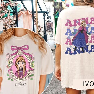 May include: Two ivory t-shirts with different designs featuring the character Anna from the movie Frozen. One shirt has a graphic of Anna in a pink frame with flowers and pearls. The other shirt has a graphic of Anna with the word "Anna" repeated four times in a retro font.