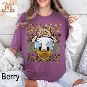 May include: Purple t-shirt with a graphic of Daisy Duck wearing a safari hat and surrounded by animal print designs. The text "Daisy Day Daisy" is printed on the shirt.