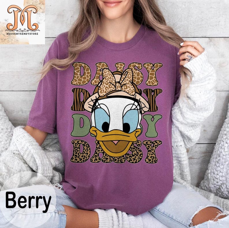 May include: Purple t-shirt with a graphic of Daisy Duck wearing a safari hat and surrounded by animal print designs. The text "Daisy Day Daisy" is printed on the shirt.