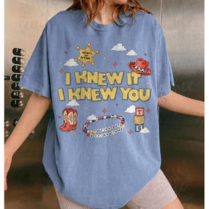 May include: A light blue short-sleeved t-shirt featuring a graphic print with the text I KNEW IT I KNEW YOU. The design includes a yellow star, a red cowboy hat, red cowboy boots, a beaded bracelet with the text YOU'VE GOT A FRIEND IN ME, and blocks.