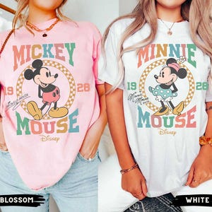 May include: Two t-shirts with a retro design featuring Mickey Mouse and Minnie Mouse. The Mickey Mouse shirt is pink with the text "Mickey Mouse" and "Disney" in a vintage font. The Minnie Mouse shirt is white with the text "Minnie Mouse" and "Disney" in a vintage font.