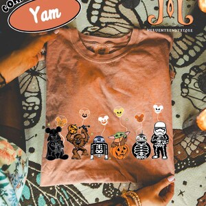 Op de afbeelding: Een Comfort Colors "Yam" t-shirt met een afbeelding van Star Wars-personages met Halloween-ballonnen. De personages zijn Darth Vader, Chewbacca, R2-D2, Baby Yoda, een pompoen, een skelet en een Stormtrooper.