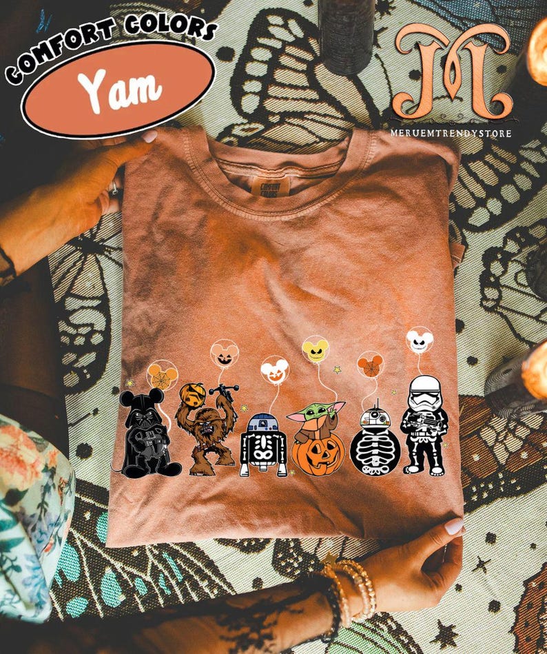 Op de afbeelding: Een Comfort Colors "Yam" t-shirt met een afbeelding van Star Wars-personages met Halloween-ballonnen. De personages zijn Darth Vader, Chewbacca, R2-D2, Baby Yoda, een pompoen, een skelet en een Stormtrooper.