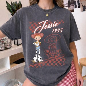K&ouml;nnte beinhalten: Dunkelgraues T-Shirt mit einer Grafik der Figur Jessie aus Toy Story. Das Design enth&auml;lt den Text "Jessie 1995" und ein rot-wei&szlig; kariertes Muster. Das T-Shirt hat kurze &Auml;rmel und einen Rundhalsausschnitt.