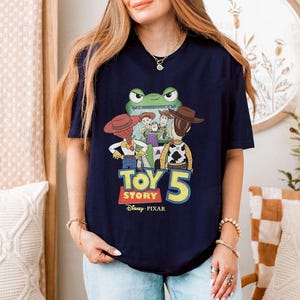 May include: Navy blue t-shirt featuring a colorful graphic of characters from the Toy Story franchise, including Woody, Buzz Lightyear, and Jessie. The text "Toy Story 5" is prominently displayed, along with the Disney Pixar logo.