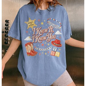 May include: A light blue short-sleeve t-shirt featuring a graphic print with the text I KNEW IT, I KNEW YOU, TOY STORY, INFINITY AND BEYOND, and YOU'VE GOT A FRIEND IN ME. The design includes a gold sheriff star, cowboy boots, a hat, and blocks.