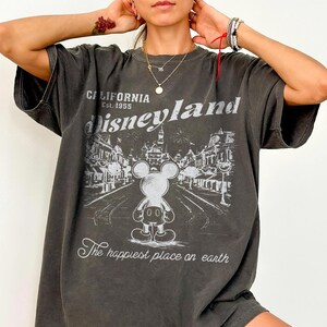 以下が含まれることがあります： ディズニーランドのグラフィックがプリントされた濃いグレーのTシャツ。「California Est. 1955 Disneyland」の文字とミッキーマウスのイラスト、パークの風景が描かれています。「The happiest place on earth」の文字が下にあります。