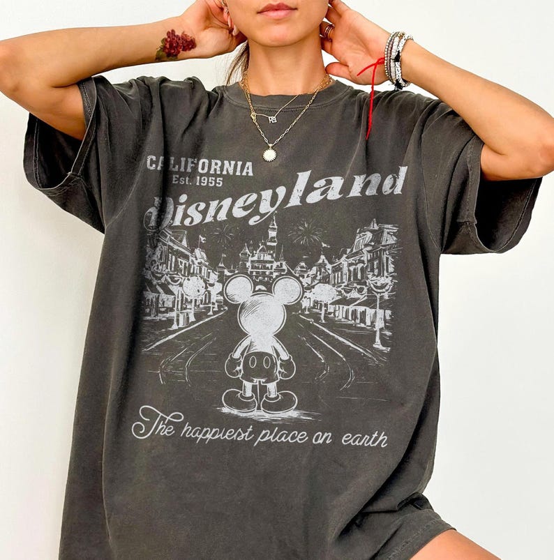 以下が含まれることがあります： ディズニーランドのグラフィックがプリントされた濃いグレーのTシャツ。「California Est. 1955 Disneyland」の文字とミッキーマウスのイラスト、パークの風景が描かれています。「The happiest place on earth」の文字が下にあります。