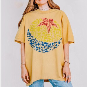 May include: Mustard yellow t-shirt featuring a large graphic of a red star, yellow circle, and blue crescent moon, all filled with various shapes. The shirt has short sleeves and a crew neck.