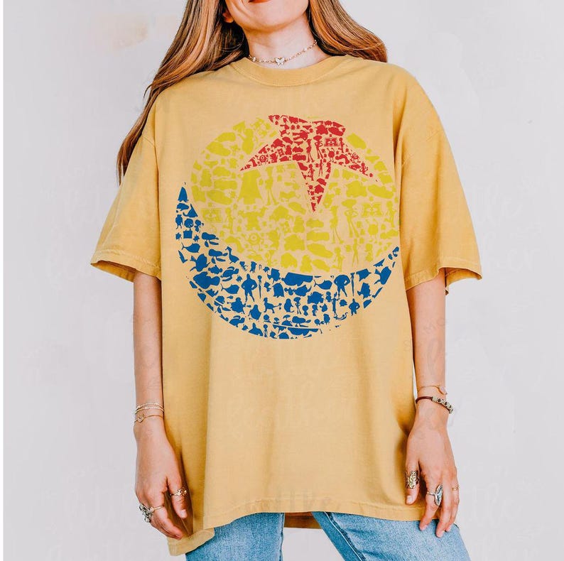 May include: Mustard yellow t-shirt featuring a large graphic of a red star, yellow circle, and blue crescent moon, all filled with various shapes. The shirt has short sleeves and a crew neck.