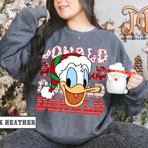 May include: Dark heather gray sweatshirt featuring a Donald Duck graphic wearing a Santa hat. The text "DONALD DAD" is displayed in a festive font. A Santa mug is held in the hand. The words "DARK HEATHER" are at the bottom.