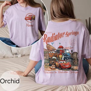 Op de afbeelding: Orchidee-kleurig T-shirt met een afbeelding van een rode raceauto en de tekst "Name" op de voorkant. De achterkant van het shirt toont een kleurrijke illustratie van personages en de tekst "Radiator Springs, California".