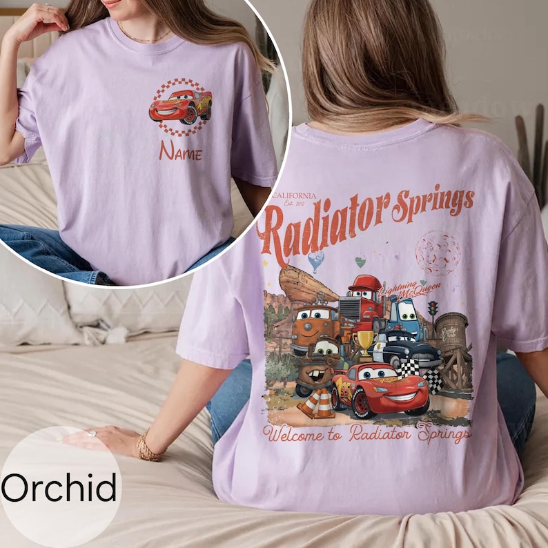 Op de afbeelding: Orchidee-kleurig T-shirt met een afbeelding van een rode raceauto en de tekst "Name" op de voorkant. De achterkant van het shirt toont een kleurrijke illustratie van personages en de tekst "Radiator Springs, California".