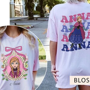 May include: Two pink t-shirts with a Frozen character design. One shirt features a portrait of Anna in a frame with flowers and the text "Anna". The other shirt has the text "Anna" repeated in a retro style with a cartoon image of Anna in a blue and white dress.