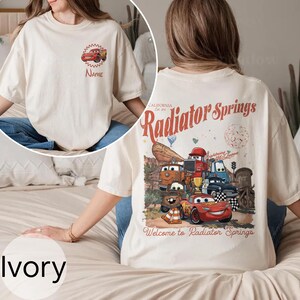 Op de afbeelding: Ivoor kleurig T-shirt met een cartoon auto ontwerp op de voorkant en een grotere afbeelding op de achterkant. De afbeelding op de achterkant bevat de tekst "Radiator Springs" en diverse auto characters. Het shirt is gemaakt van zacht materiaal.