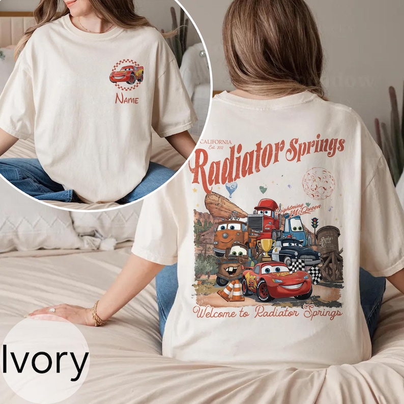 Op de afbeelding: Ivoor kleurig T-shirt met een cartoon auto ontwerp op de voorkant en een grotere afbeelding op de achterkant. De afbeelding op de achterkant bevat de tekst "Radiator Springs" en diverse auto characters. Het shirt is gemaakt van zacht materiaal.