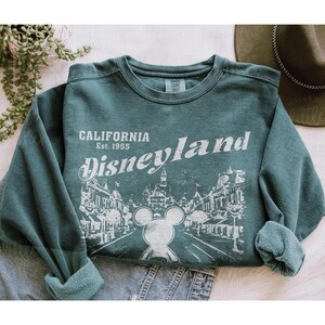 以下が含まれることがあります： 白文字で「CALIFORNIA Disneyland Est. 1955」と書かれた、ティール色のスウェットシャツ。ミッキーマウスとディズニーランドのイラストが描かれています。スウェットシャツは袖をまくっています。