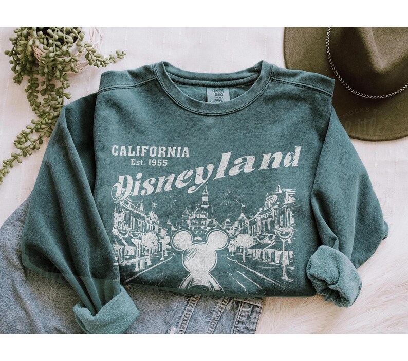 以下が含まれることがあります： 白文字で「CALIFORNIA Disneyland Est. 1955」と書かれた、ティール色のスウェットシャツ。ミッキーマウスとディズニーランドのイラストが描かれています。スウェットシャツは袖をまくっています。