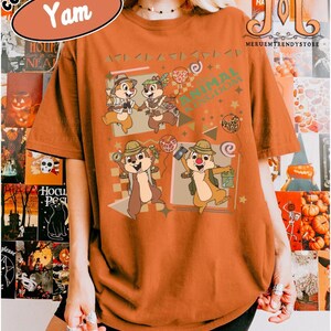 May include: A yam-colored t-shirt featuring a graphic print of four cartoon chipmunks dressed in safari gear. The design includes the text Animal Kingdom, various geometric shapes, stars, and balloons. The shirt has short sleeves and a crew neckline.