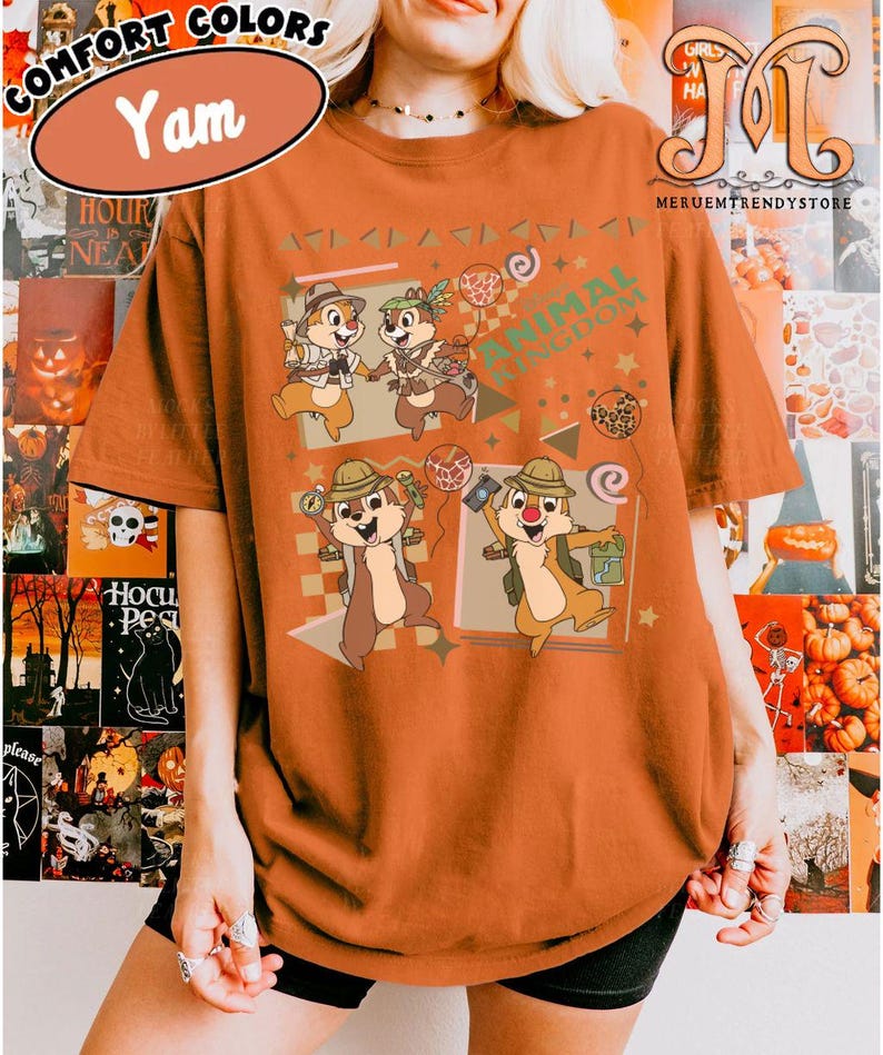 May include: A yam-colored t-shirt featuring a graphic print of four cartoon chipmunks dressed in safari gear. The design includes the text Animal Kingdom, various geometric shapes, stars, and balloons. The shirt has short sleeves and a crew neckline.