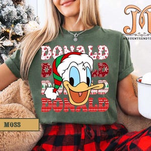 May include: Moss green t-shirt featuring a graphic of Donald Duck in a Santa hat with the text "DONALD" in a festive font. The shirt is paired with red and black plaid pants, creating a holiday theme.