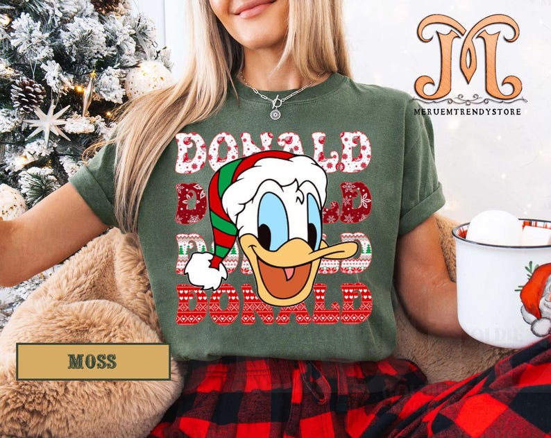 May include: Moss green t-shirt featuring a graphic of Donald Duck in a Santa hat with the text "DONALD" in a festive font. The shirt is paired with red and black plaid pants, creating a holiday theme.