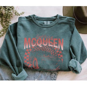 May include: A teal-colored sweatshirt with the text "Lightning McQueen" in a gradient red font. Below the text is a red and white checkered design with a car wheel. The sweatshirt has rolled-up sleeves and a crew neck.