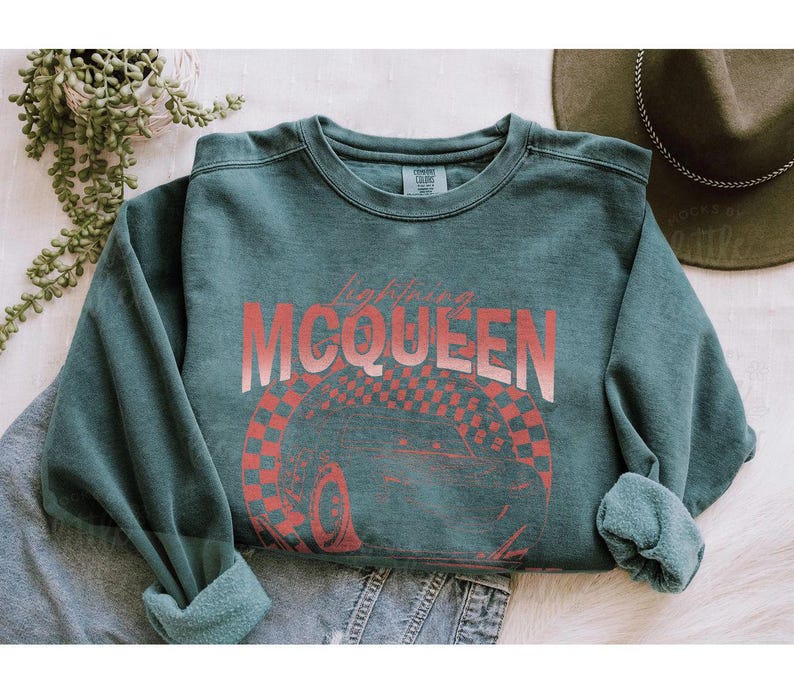 May include: A teal-colored sweatshirt with the text "Lightning McQueen" in a gradient red font. Below the text is a red and white checkered design with a car wheel. The sweatshirt has rolled-up sleeves and a crew neck.