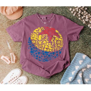 May include: A mauve-colored t-shirt featuring a large graphic design. The design is a circle divided into three sections: a red star, yellow shapes, and blue shapes. The shirt is surrounded by accessories.