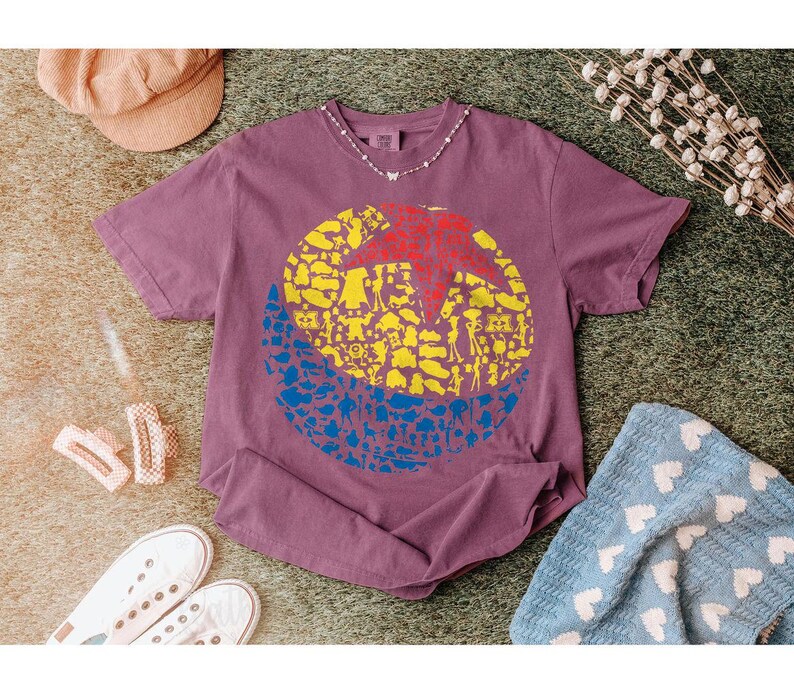 May include: A mauve-colored t-shirt featuring a large graphic design. The design is a circle divided into three sections: a red star, yellow shapes, and blue shapes. The shirt is surrounded by accessories.