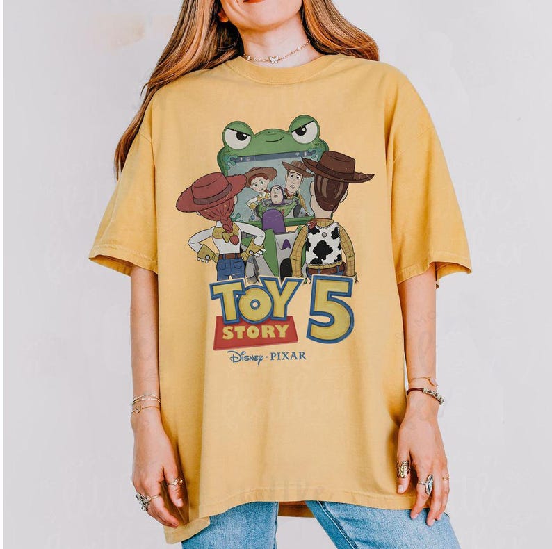May include: Mustard yellow t-shirt featuring a graphic from the Disney Pixar movie Toy Story 5. The design includes characters like Woody, Buzz Lightyear, and others, with the movie title and logo prominently displayed.