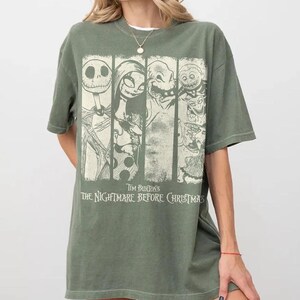 以下が含まれることがあります： オリーブグリーンの半袖Tシャツ。ティム・バートンの『ナイトメアー・ビフォア・クリスマス』のキャラクターがクリーム色のグラフィックでプリントされています。デザインは4つの縦パネルに分かれており、ジャック・スケリントン、サリーなどが描かれています。