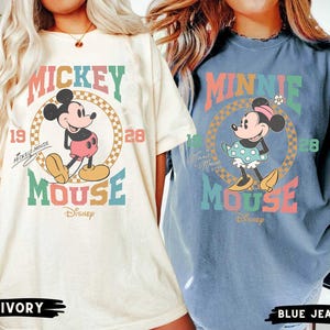 May include: Two oversized t-shirts with a vintage Mickey Mouse and Minnie Mouse design. The Mickey Mouse shirt is ivory with a red, yellow, blue, and green color scheme. The Minnie Mouse shirt is blue jean with a red, yellow, blue, and green color scheme. Both shirts feature the text "Mickey Mouse" and "Minnie Mouse" with the word "Mouse" in a rainbow color scheme.