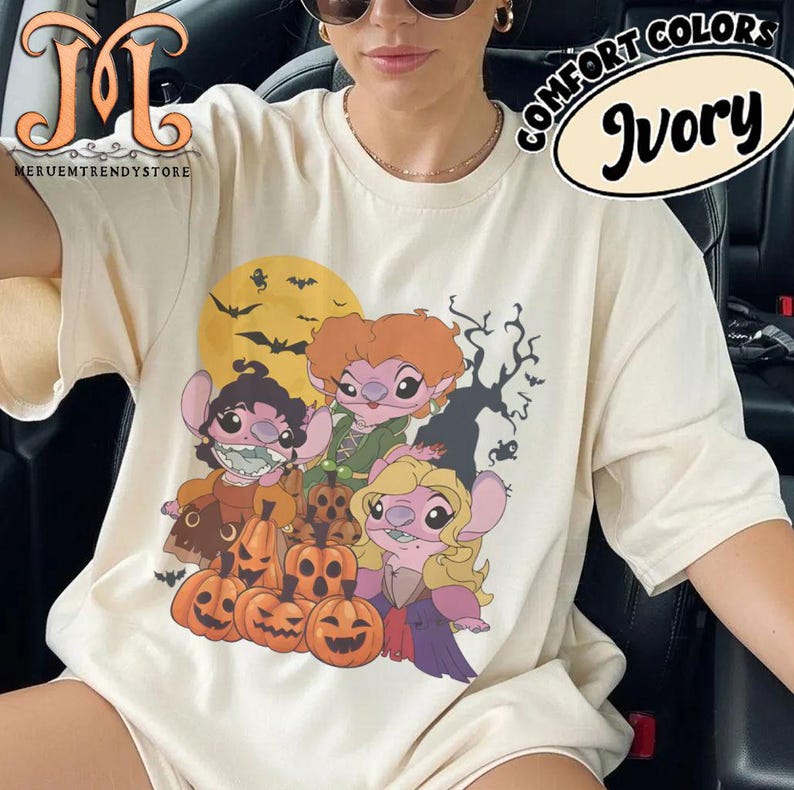 May include: Ivory-colored t-shirt featuring a Halloween-themed graphic. The design includes cartoon characters, pumpkins, bats, and a full moon. The text "COMFORT COLORS" and "Ivory" are printed on the shirt. The brand name "MERUEMTRENDYSTORE" is also visible.