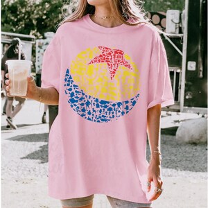 May include: A light pink t-shirt featuring a colorful graphic design. The design includes a yellow circle with a red star, and a blue crescent shape filled with various small figures. The shirt is a casual, oversized style.