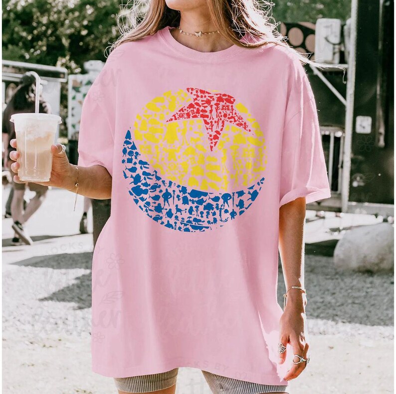 May include: A light pink t-shirt featuring a colorful graphic design. The design includes a yellow circle with a red star, and a blue crescent shape filled with various small figures. The shirt is a casual, oversized style.