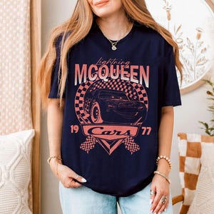 May include: Navy blue t-shirt featuring a graphic of Lightning McQueen from the Cars movie. The design includes the text "Lightning McQueen," "Cars," and the numbers "19" and "77" in a retro style. The graphic is in shades of pink and white.