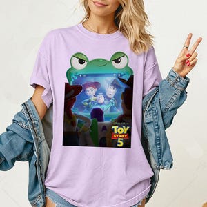 May include: A lilac t-shirt with a green frog-shaped screen showing a scene from "Toy Story 5." The graphic includes Woody, Buzz Lightyear, and Jessie. The top has short sleeves and a round neck.
