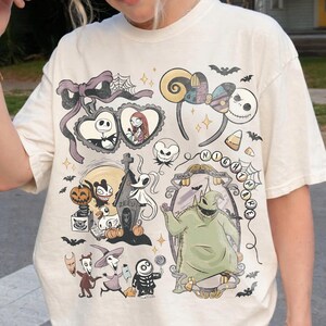 May include: An off-white t-shirt featuring a graphic print of characters and icons from The Nightmare Before Christmas, including Jack Skellington, Sally, Zero, and Oogie Boogie, alongside bats, spiderwebs, and the text Nightmare.