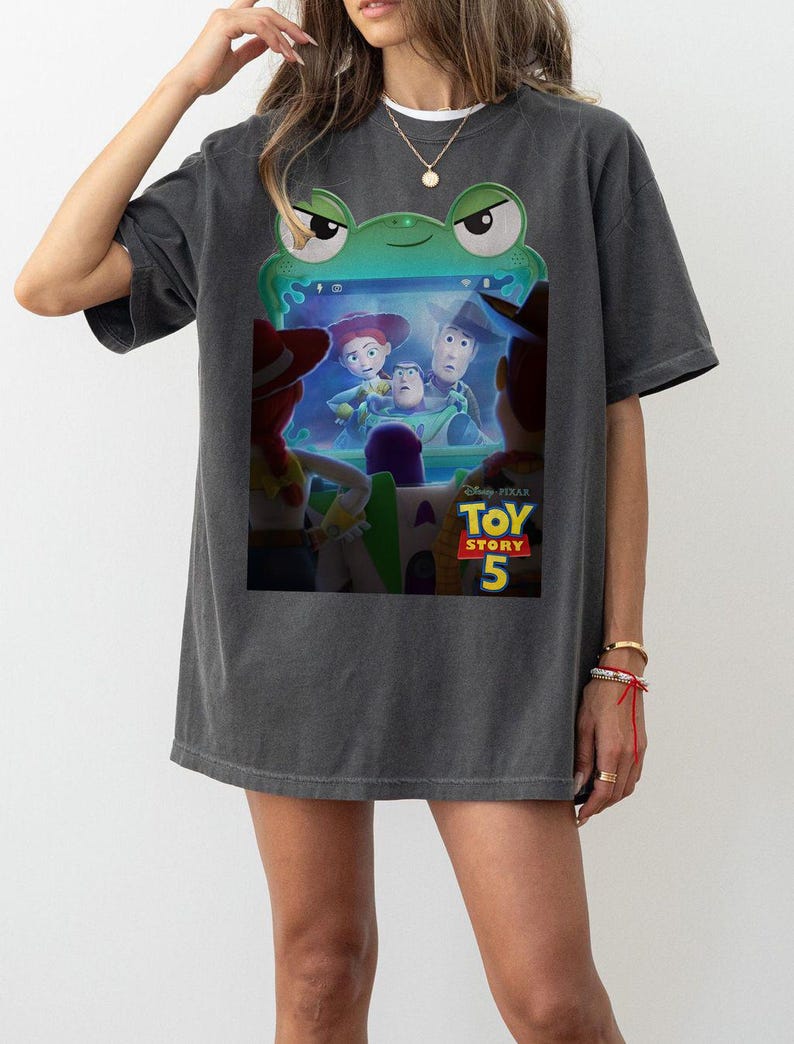 May include: Dark grey t-shirt with a green frog graphic and the text "Toy Story 5". The graphic shows characters from the animated film. The shirt has a casual, oversized fit.