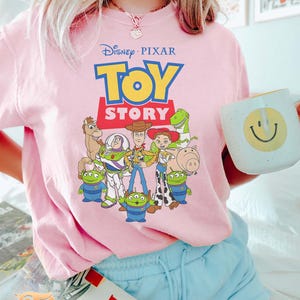 May include: A light pink t-shirt featuring the Disney Pixar Toy Story logo and characters. The design includes Woody, Buzz Lightyear, and other characters from the animated film. The person is holding a white mug with a yellow smiley face.