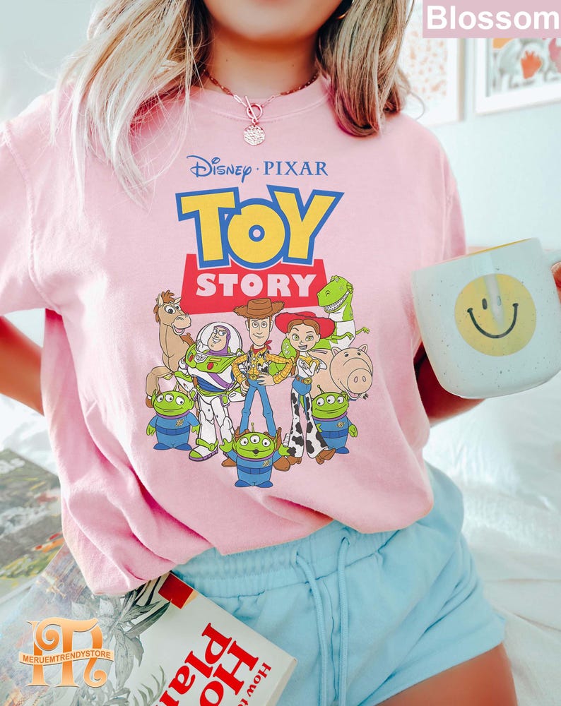 May include: A light pink t-shirt featuring the Disney Pixar Toy Story logo and characters. The design includes Woody, Buzz Lightyear, and other characters from the animated film. The person is holding a white mug with a yellow smiley face.