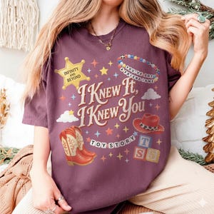 May include: A maroon short-sleeve t-shirt featuring a graphic print with the text I KNEW IT, I KNEW YOU, TOY STORY, INFINITY AND BEYOND, and YOU'VE GOT A FRIEND IN ME. The design includes a yellow sheriff star, cowboy boots, a hat, and blocks.