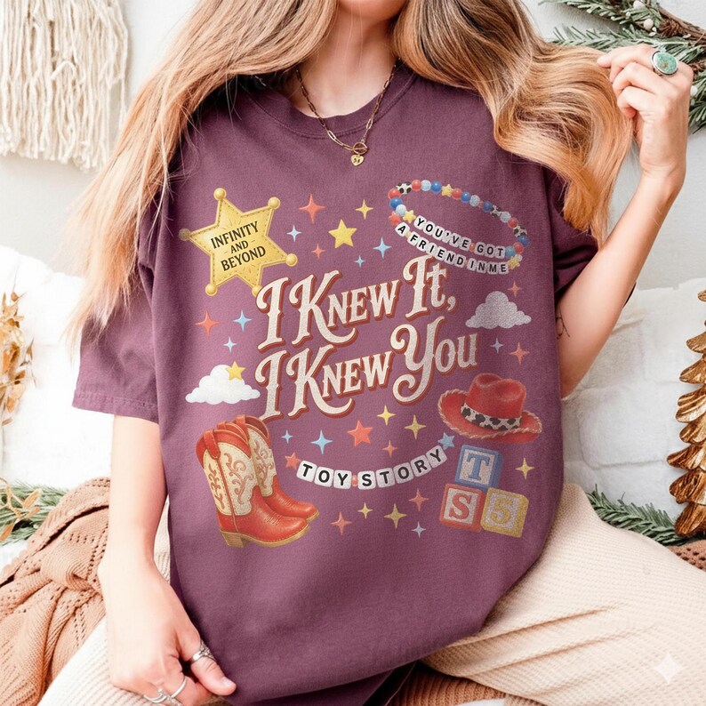 May include: A maroon short-sleeve t-shirt featuring a graphic print with the text I KNEW IT, I KNEW YOU, TOY STORY, INFINITY AND BEYOND, and YOU'VE GOT A FRIEND IN ME. The design includes a yellow sheriff star, cowboy boots, a hat, and blocks.