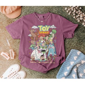 Puede incluir: Una camiseta de color malva con un gr&aacute;fico de personajes de la pel&iacute;cula animada "Toy Story". El dise&ntilde;o incluye a Woody, Buzz Lightyear y otros personajes, con el texto "TOY STORY" y "YOU'VE GOT A FRIEND IN ME."