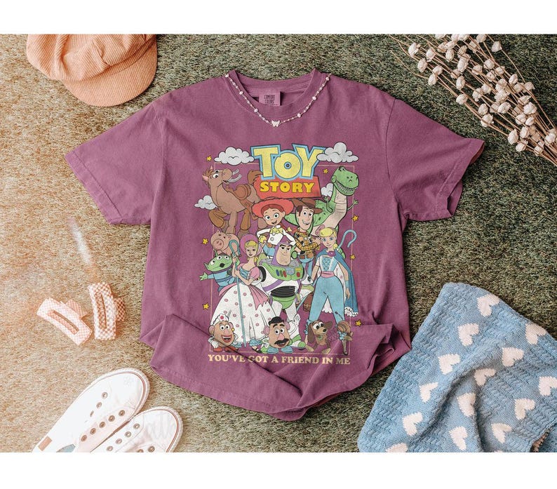 Puede incluir: Una camiseta de color malva con un gr&aacute;fico de personajes de la pel&iacute;cula animada "Toy Story". El dise&ntilde;o incluye a Woody, Buzz Lightyear y otros personajes, con el texto "TOY STORY" y "YOU'VE GOT A FRIEND IN ME."