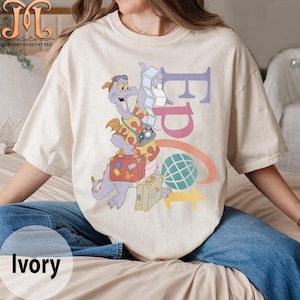 May include: A cream colored t-shirt with a cartoon purple dragon wearing a Hawaiian shirt and carrying luggage. The dragon is standing in front of the word "Epcot" in rainbow colors.